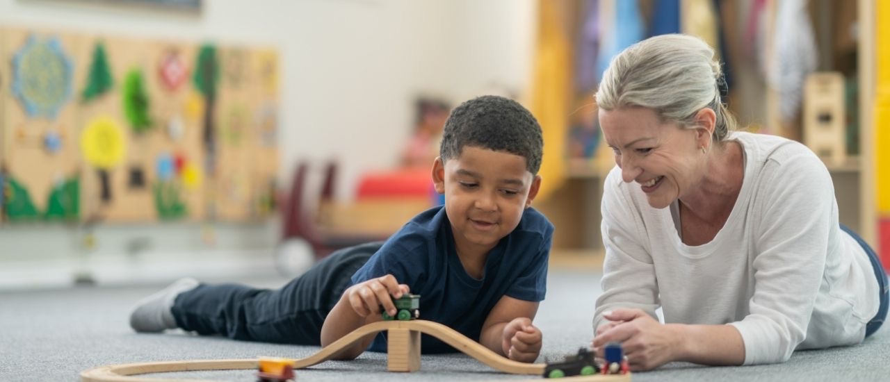 A therapist and a young boy smiling as they build a wooden train track on the floor